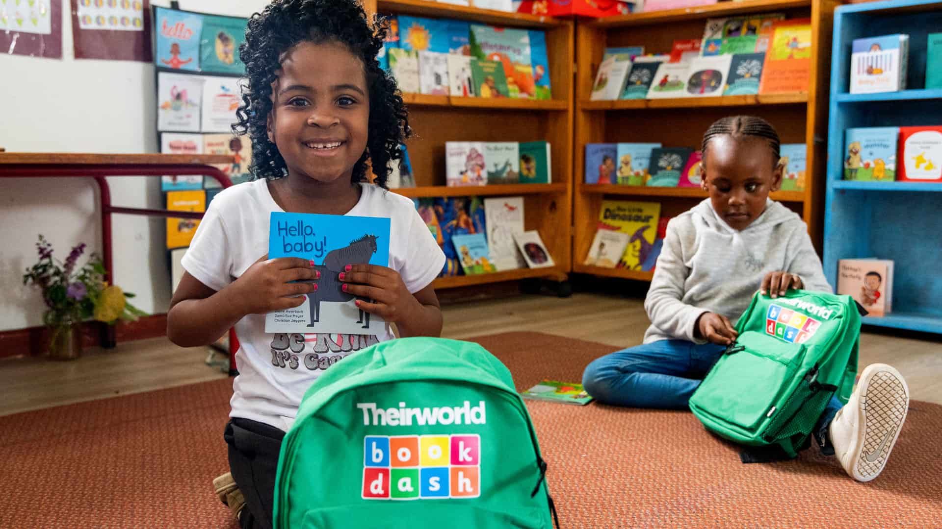 Two children sitting on the floor with backpacks in front of bookshelves.