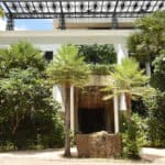 Front view of a modern three-story building surrounded by lush greenery and tropical plants. The white facade is accented with large windows and a black lattice rooftop structure. A large stone block is placed in front of the entrance.