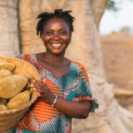 A woman wearing a colorful Aduna patterned dress stands outdoors in front of a large tree, smiling and holding a basket filled with large, oval-shaped fruits. The background is a dry, earthy landscape with additional trees visible in the distance.