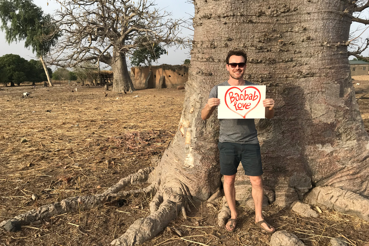 Andrew Hunt standing in front of a large baobab tree holds a sign with "Baobab Love" written inside a heart. Sporting sunglasses, a gray t-shirt, and shorts branded with Aduna, he poses against a backdrop of more baobab trees, a small building, and an open, arid landscape.