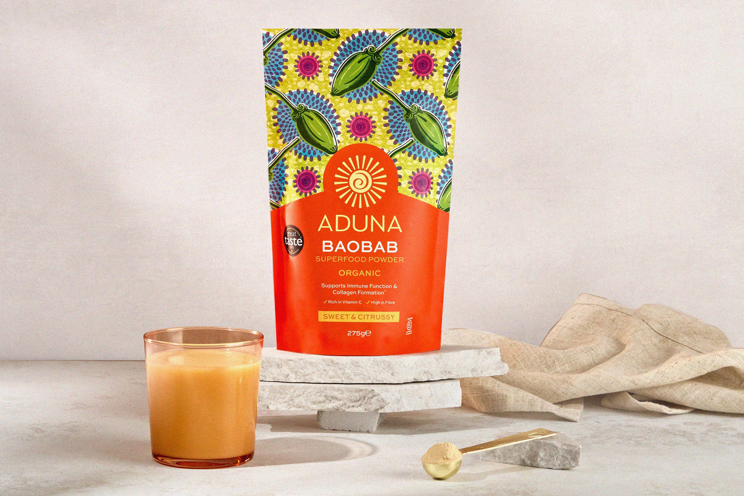 A colorful packet of Aduna Baobab Superfood Powder stands on stacked stone slabs. In front of it, a glass of yellow-orange drink, a scoop of powder, and a gold spoon are placed on a white surface. The Aduna packet features a bright design with blue, green, and yellow patterns.