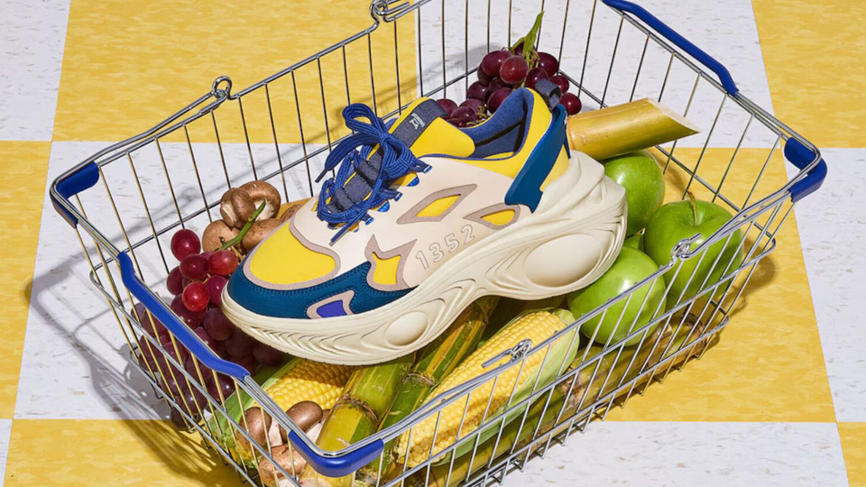 A shopping basket filled with various fruits and vegetables, including green apples, bananas, corn, and grapes, also contains a trendy sneaker with a distinctive blue, yellow, and beige design. The basket is placed on a yellow and white checkered floor, resembling an organized directory of colorful items.