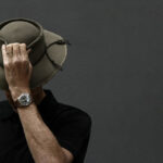 Scott Poynton person wearing a dark shirt, tilting a wide-brimmed hat down to cover their face, stands against a plain, dark background. They have a wristwatch and a ring on their left hand, embodying the essence of global citizenship through their timeless yet modern style.
