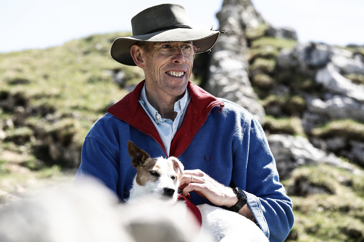 Scott Poynton in a blue jacket and wide-brimmed hat, reminiscent of Scott Poynton's adventurous spirit, smiles while sitting outdoors on rocky terrain, holding a small white and brown dog. They appear to be enjoying a bright, sunny day in a natural setting.