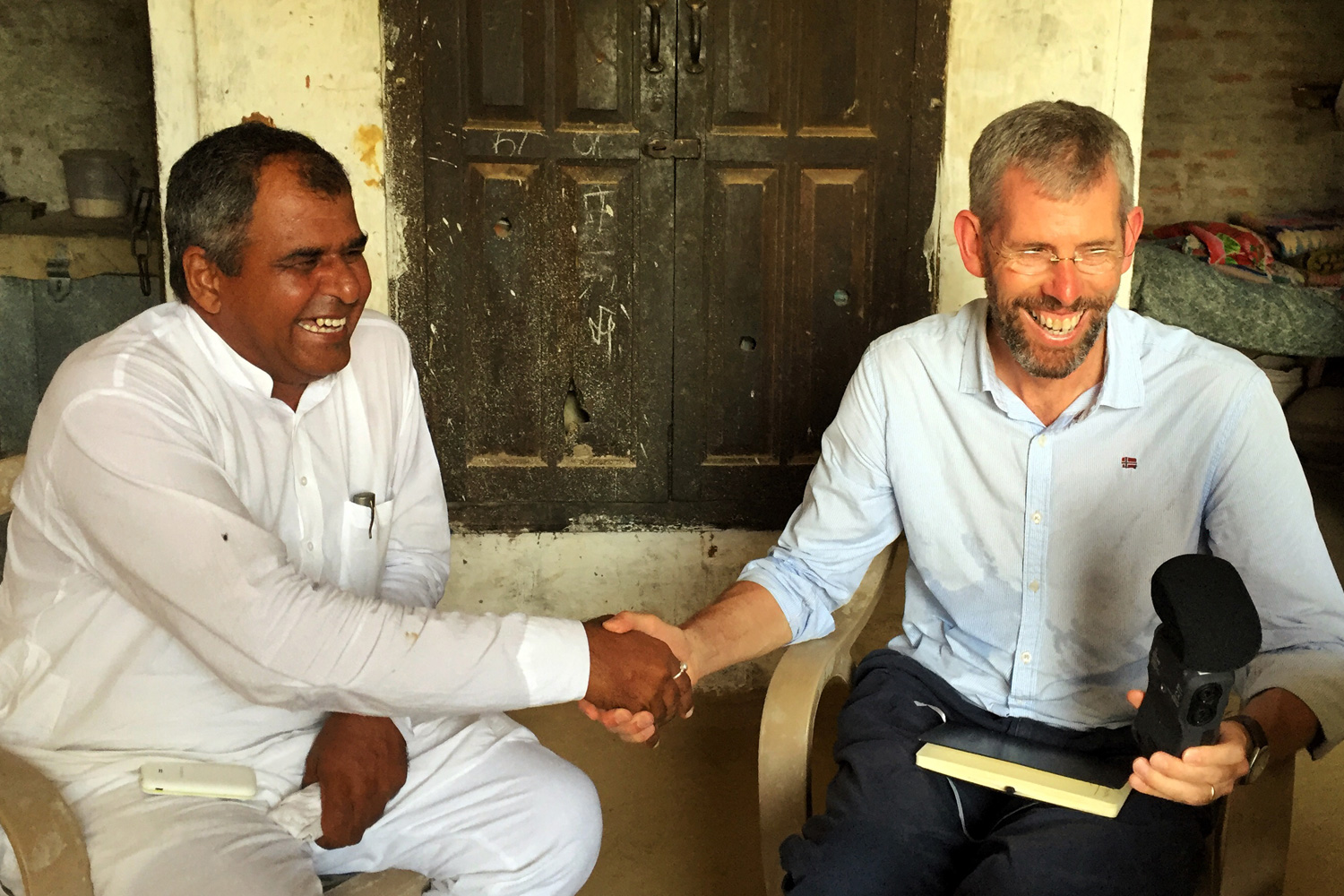 Two men are seated, shaking hands and smiling. One man, dressed in white traditional clothing, holds a small notebook, while the other man, identified as Scott Poynton and wearing glasses and a light blue shirt, holds a recorder. They are sitting in front of a dark wooden door and seem to be in a warm, friendly setting.