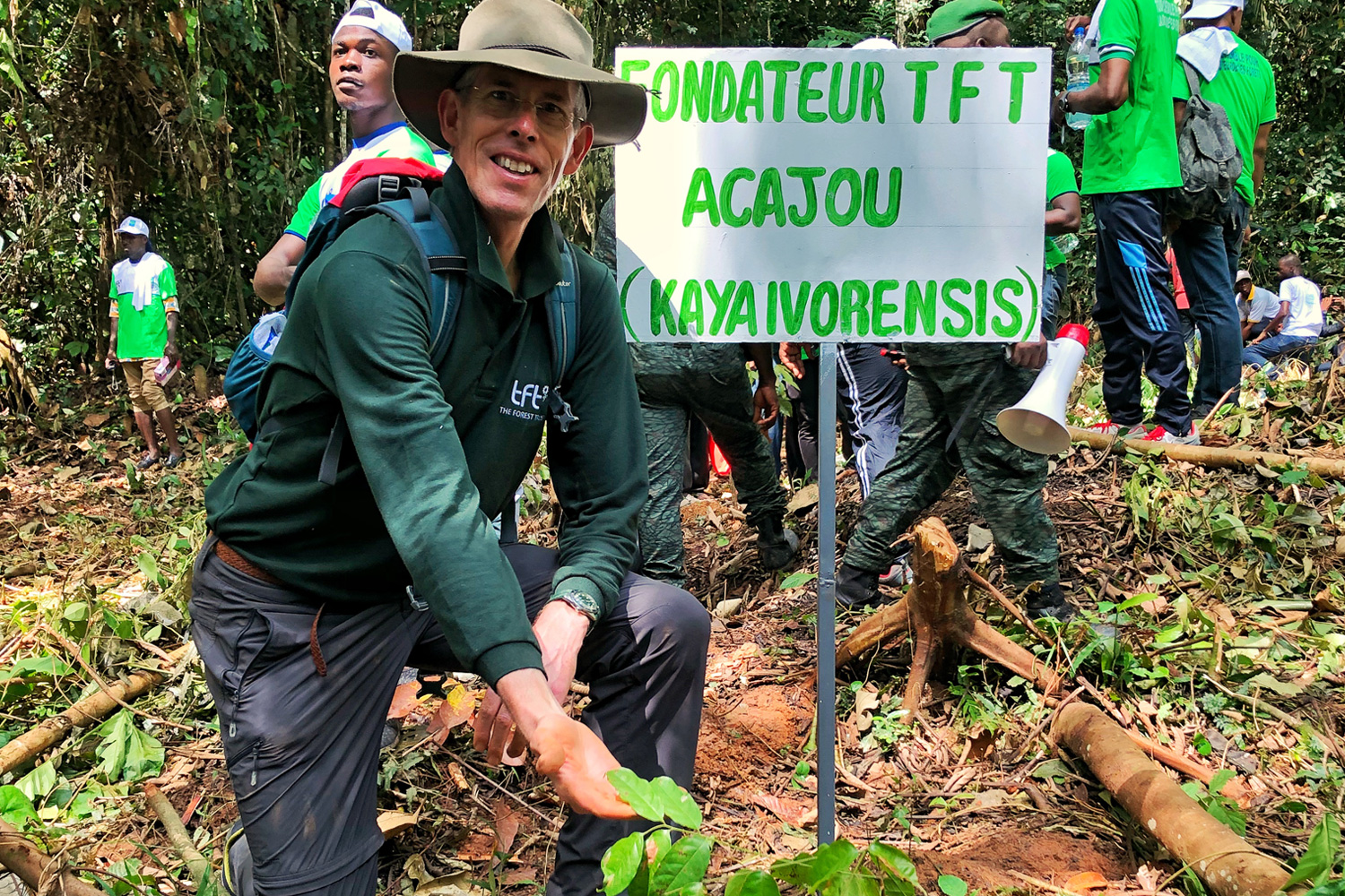 Scott Poynton wearing a hat and green shirt kneels next to a sign in a forested area. The sign reads "FONDATEUR TFT ACAJOU (KAYAIVORENSIS)" in green and white. Scott Poynton can be seen in the background, along with others in green shirts, engaging in an outdoor activity.