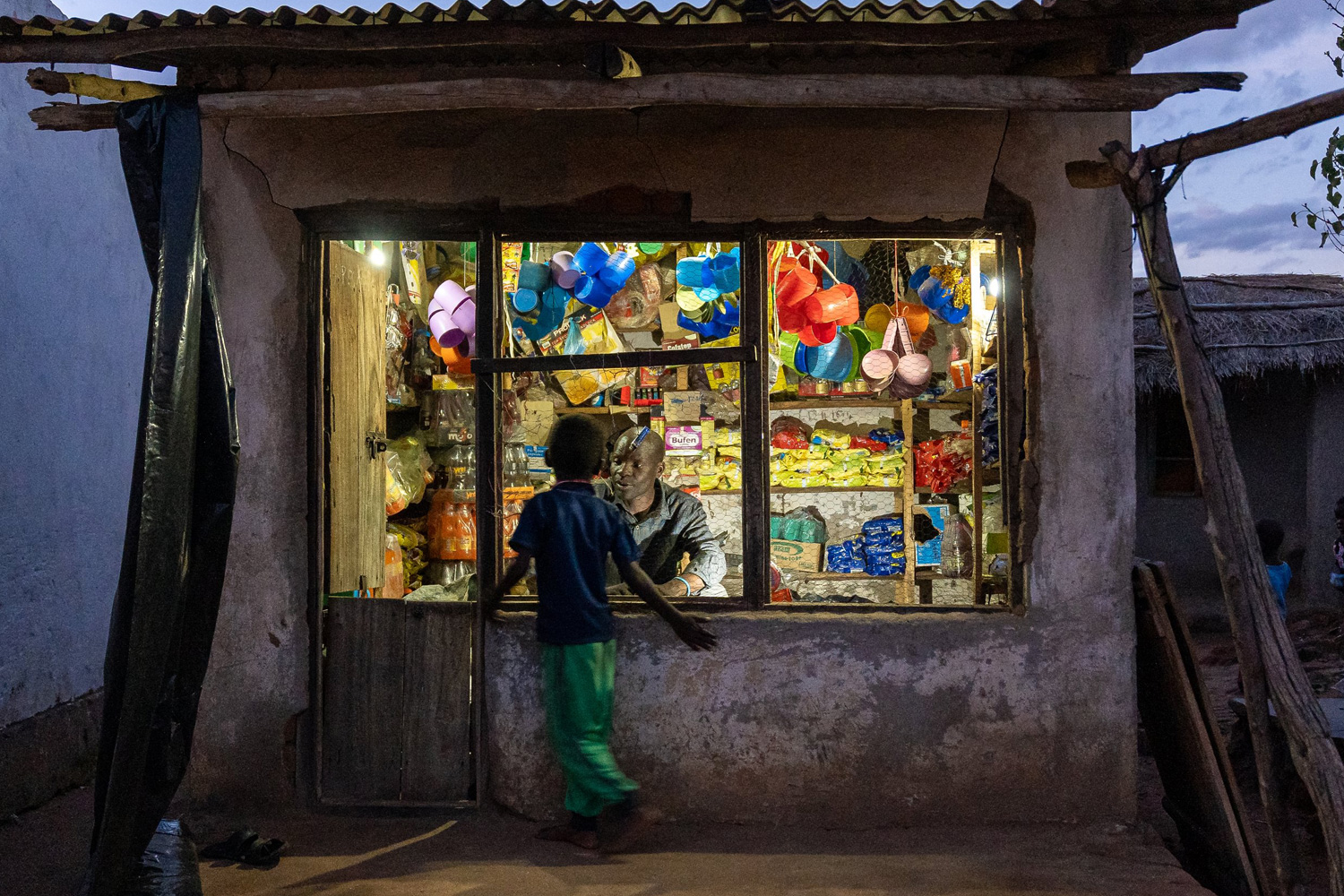 In the dim evening light, a small shop, powered by discrete solar panels, lit from within, displays various colorful goods. A child stands at the entrance, seemingly conversing with the shopkeeper. The shop's rustic exterior contrasts with the bright, vibrant items visible through the window.