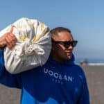 A person wearing sunglasses and a blue hoodie with "UOCEAN 2050" printed on it stands on a beach, representing La Isla Network, carrying a white sack over their shoulder. The ocean is visible in the background under a clear sky.