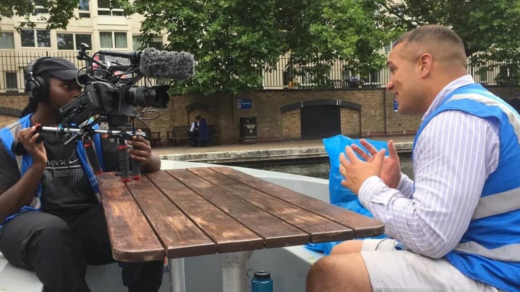 A man is being interviewed by someone holding a video camera on a wooden table in a boat for La Isla Network. The interviewer is wearing a headset and has a microphone attached to the camera. The background features a canal, trees, and a brick building.