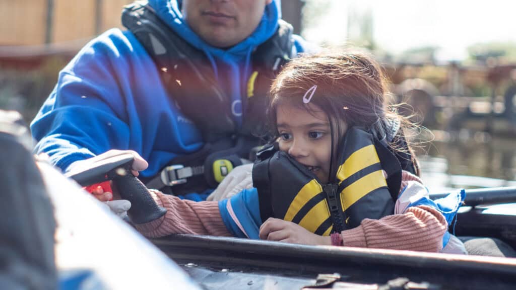 A young child wearing a yellow and black life jacket sits in a boat, closely supervised by an adult in a blue life jacket. The child is holding a boat control lever with a focused expression, as the water reflects sunlight in the background, enjoying an adventure shared through La Isla Network.