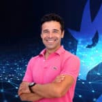 A beVisioneers man, donning a pink polo shirt, smiles confidently with his arms crossed. Behind him, a dark background showcases abstract blue network lines and a large animal-like silhouette. His dark hair complements his poised demeanor perfectly.