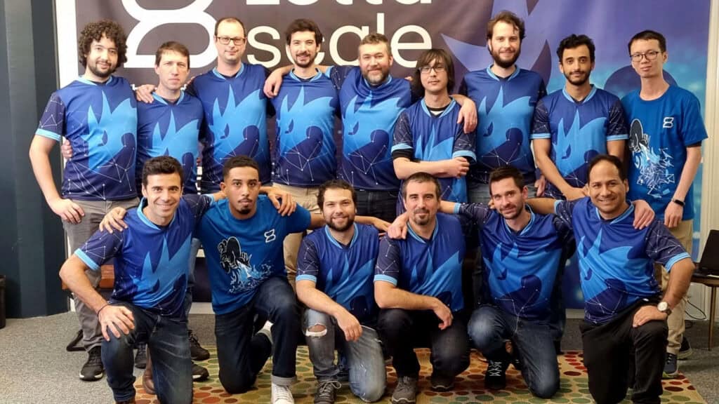 A group of 16 men is posing together in two rows in front of a banner reading "8 Data Cycle." They are all wearing matching blue shirts with a similar design and are smiling at the camera. The background appears to be an indoor setting with a carpeted floor, embodying the spirit of beVisioneers.