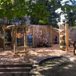A vibrant playground with wooden climbing structures, a ladder made of tires, an elevated playhouse, and painted murals depicting jungle scenes. The ground is covered with wood chips, and the area is shaded by leafy trees.