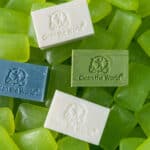 A collection of green soap bars with four different soaps on top, each imprinted with "Clean the World" and a recycling symbol. The soaps are white, beige, blue, and green, contrasting with the uniform green bars beneath.