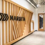 A modern office hallway with a wooden slatted wall feature displaying the name "Marqeta," championing financial inclusion. A person walks down the corridor towards a door, and the ceiling showcases exposed industrial-style elements.